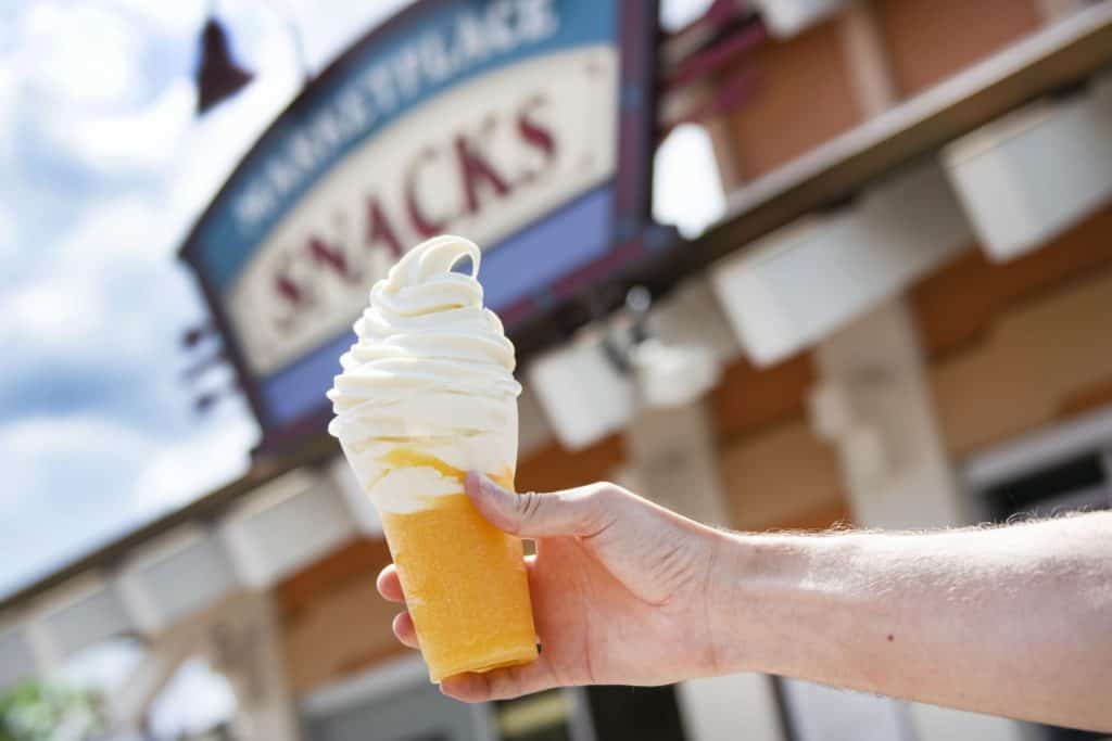 A swirl of creamy soft serve ice cream sits in a cup, channeling the classic Vanilla Orange Slushy Float from Disney Springs, with a blurred Disney sign creating magical vibes in the background.