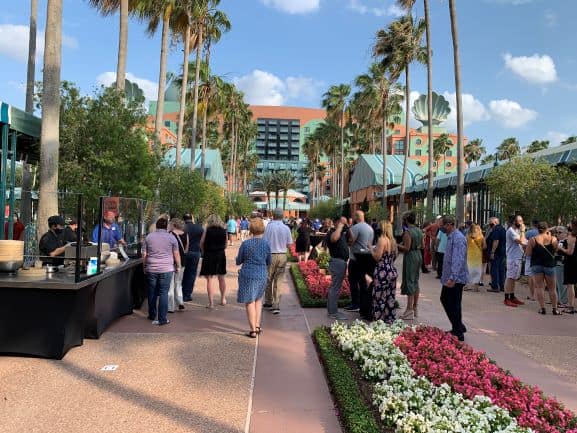 Fans enjoy the vibrant atmosphere of the Swan and Dolphin Spring Food & Wine Classic at Walt Disney World, mingling among palm trees and colorful flowerbeds outside the iconic resort under a bright blue sky dotted with clouds.