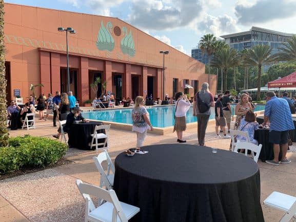 Guests relax around outdoor tables and lounge by the shimmering pool in front of the iconic Swan and Dolphin Resort, surrounded by swaying palm trees under clear blue skies. The vibrant scene buzzes with excitement during the Spring Food & Wine Classic at Walt Disney World, as attendees savor culinary delights and festive entertainment.