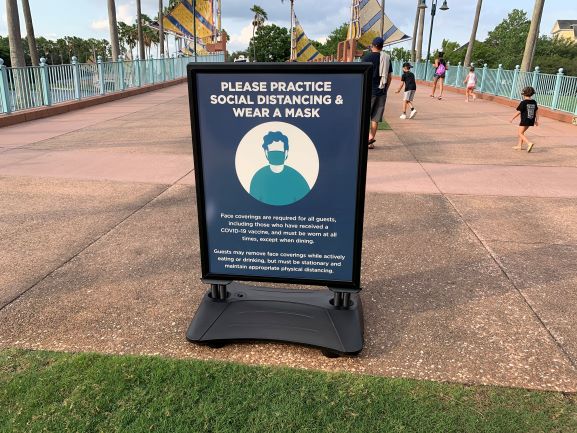 A sign on the bridge at Disney’s Swan and Dolphin Resort reminds guests to wear masks and keep a safe distance; fellow visitors stroll in the background, likely on their way to experience the Spring Food & Wine Classic festivities.