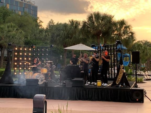 A live band entertains guests on an outdoor stage at sunset, with iconic palm trees and whimsical playground structures in the background, creating a festive vibe for the Spring Food & Wine Classic at Walt Disney World Swan and Dolphin Resort.