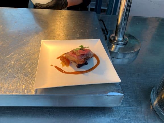A gourmet bite of meat topped with sauce, elegantly presented on a white square plate and set on a metallic counter—just the kind of culinary creation you can savor at the Spring Food & Wine Classic at Disney’s Swan and Dolphin Resort.