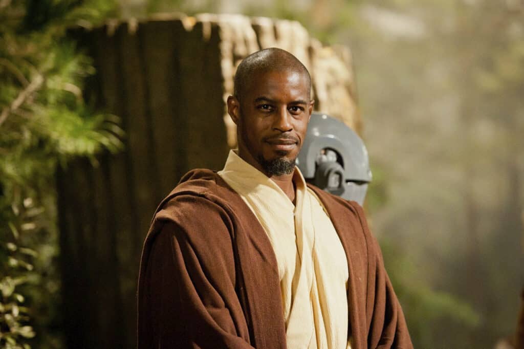 A Jedi character in a brown robe and beige tunic greets guests outdoors in a lush, wooded area at a Star Wars-themed attraction, evoking the immersive universe where characters like Jar Jar Binks come to life.