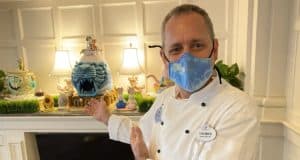 A Disney Cast Member chef in a blue mask welcomes guests to a stunning mantel display featuring elaborate cakes and beautifully crafted Easter Eggs, showcasing the magic of Disney’s culinary creations.