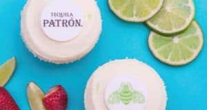 Limited-time Patrón Margarita Cupcakes, topped with white frosting and festive Disney Springs-themed toppers, sit surrounded by fresh limes and strawberries on a vibrant blue surface—an exclusive dessert inspired by the lively flavors of Disney Springs.