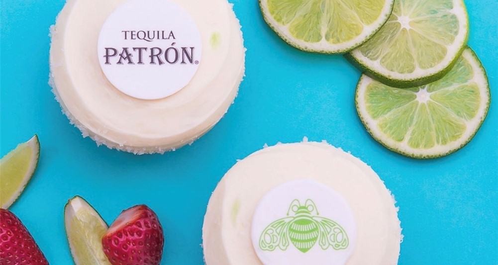 Limited-time Patrón Margarita Cupcakes, topped with white frosting and festive Disney Springs-themed toppers, sit surrounded by fresh limes and strawberries on a vibrant blue surface—an exclusive dessert inspired by the lively flavors of Disney Springs.