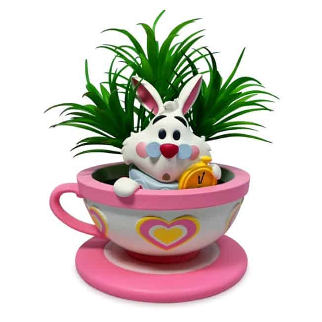 The White Rabbit from Alice in Wonderland sits in a whimsical pink teacup planter decorated with hearts, clutching his clock, surrounded by lush greenery and playful succulents—perfect for fans of classic storybook adventures at a fantasy-themed park.