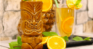 A vibrant Tiki mug with a playful Polynesian face, accompanied by a citrus-garnished orange beverage that brings to mind exclusive park refreshments, all set against the lively backdrop of fresh mint leaves—perfect for an immersive theme park tiki bar experience.