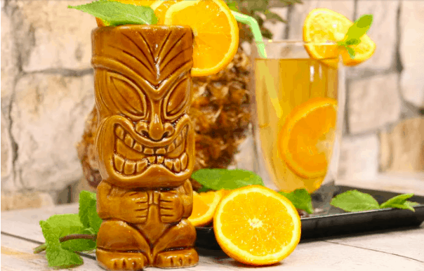 A vibrant Tiki mug with a playful Polynesian face, accompanied by a citrus-garnished orange beverage that brings to mind exclusive park refreshments, all set against the lively backdrop of fresh mint leaves—perfect for an immersive theme park tiki bar experience.