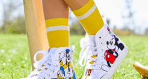 A child in bright yellow socks and playful cartoon-themed sneakers strikes a fun pose on the theme park lawn, right next to a whimsical wooden bat, ready for adventure.