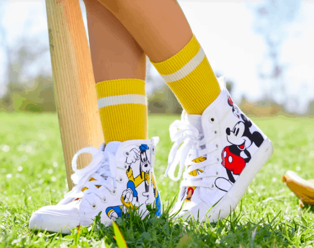 A child in bright yellow socks and playful cartoon-themed sneakers strikes a fun pose on the theme park lawn, right next to a whimsical wooden bat, ready for adventure.