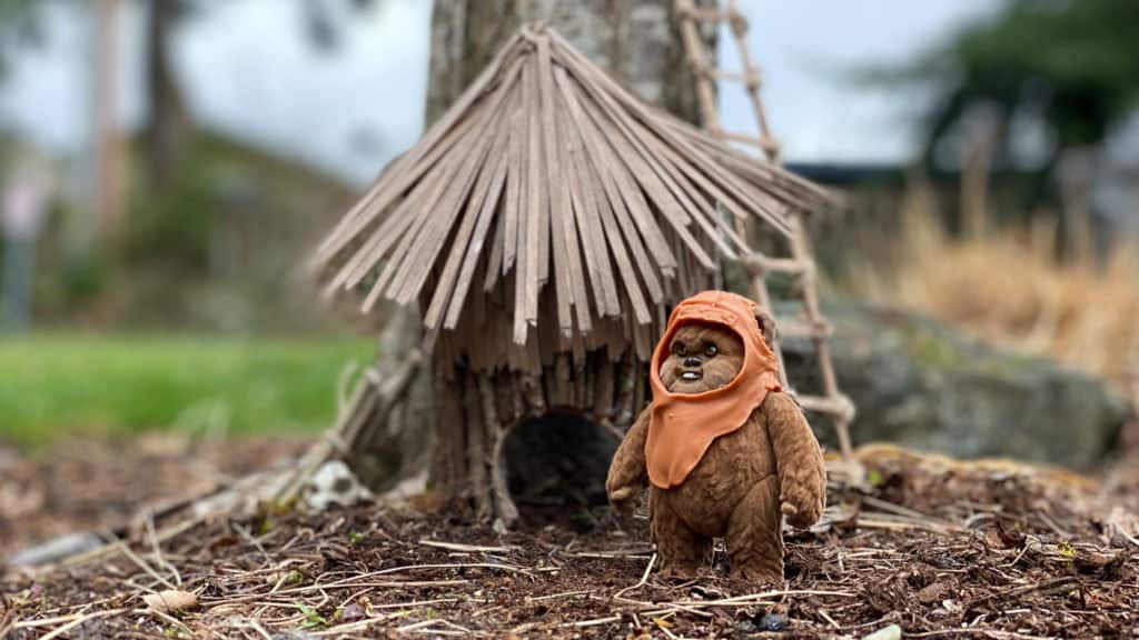 An Ewok from Star Wars stands in front of a charming miniature wooden hut at the base of a tree, evoking the magic of Endor in a whimsical fairy house display reminiscent of themed areas found at Disney parks.