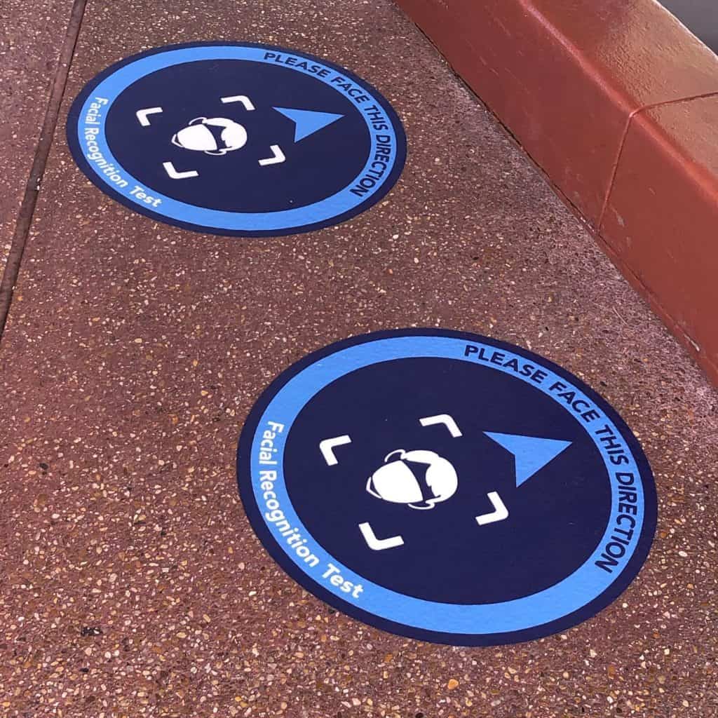 Blue floor decals inside Magic Kingdom read "Please face this direction, Facial Recognition Test" with a face icon and an arrow, helping Disney guests navigate the facial recognition check smoothly as part of their magical park entry experience.