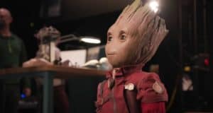 Groot, sporting a vibrant red jacket, stands indoors amidst a workbench and bustling guests in the background—capturing the excitement of meeting the lifelike Groot robot at Disney theme parks.