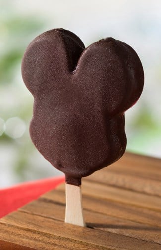 Mickey Mouse-shaped ice cream bar from a Disney collaboration series, coated in chocolate and served on a wooden stick, displayed on a rustic wooden table—just like the classic treat found at Disney Parks.