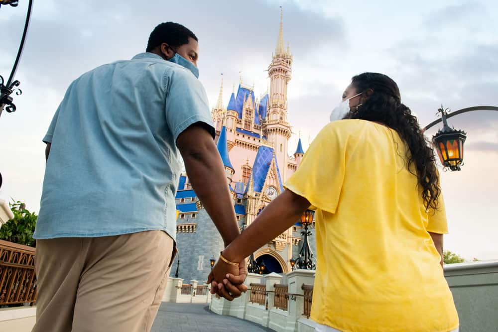 Guests holding hands and wearing masks stroll toward Cinderella Castle at Magic Kingdom during their Disney PhotoPass Session. Easily reserve your PhotoPass through the My Disney Experience App to capture every magical Disney moment.