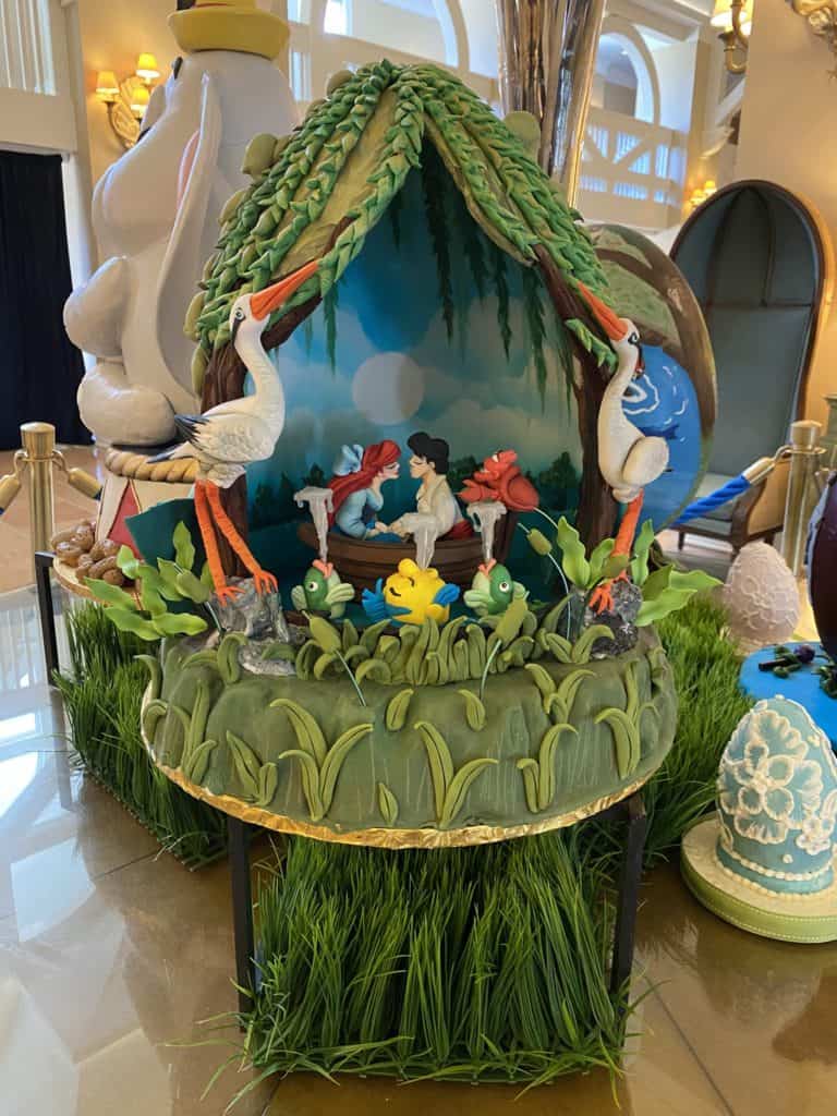 Whimsical cake featuring figurines of a man and woman, elegant swans, and playful frogs beneath enchanting trees, set against a hand-painted scenic backdrop—just like the intricate Easter Egg displays crafted by Disney’s talented Cast Members at Disney’s Yacht & Beach Club Resorts.