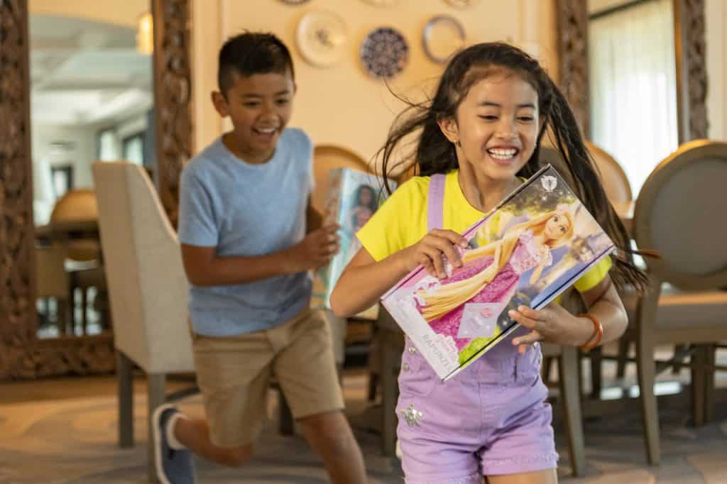 A joyful girl dashes through a vibrant, playfully decorated room, clutching her favorite classic doll as if on an imaginary adventure, while a boy follows close behind with an open storybook—capturing the energy and wonder found in theme park kids' zones.