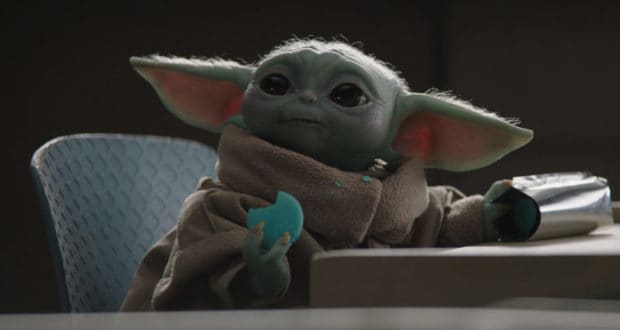 Grogu enjoys a Star Wars Day treat at Galaxy’s Edge, lounging in a chair with a blue cookie and silver snack package in hand, crumbs dotting his robe as fans celebrate around him.