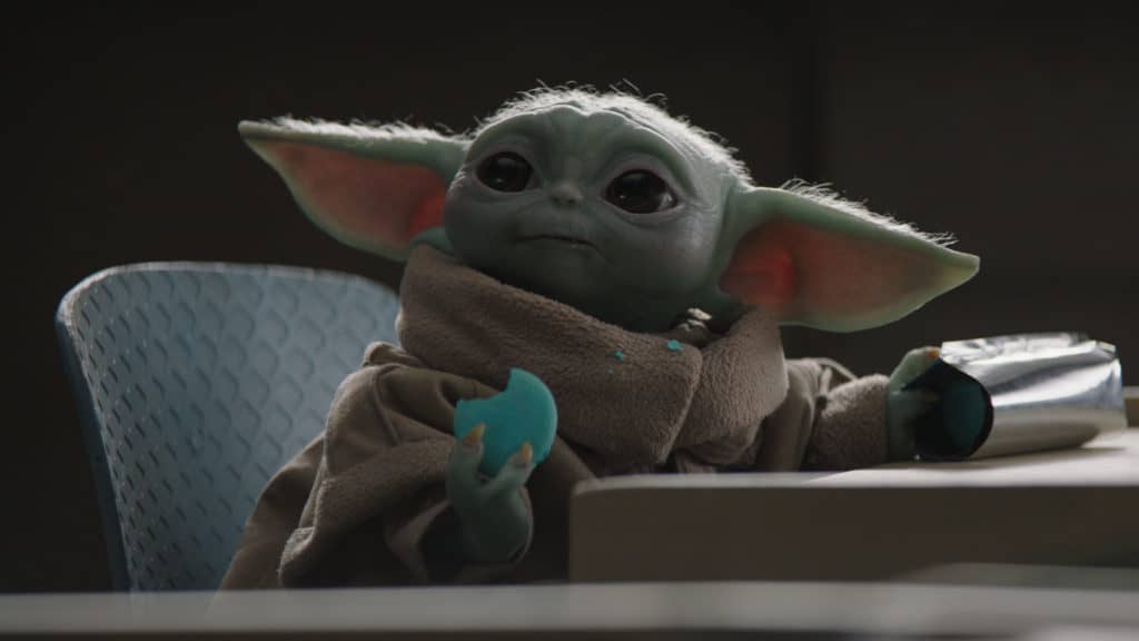 Grogu enjoys a Star Wars Day treat at Galaxy’s Edge, lounging in a chair with a blue cookie and silver snack package in hand, crumbs dotting his robe as fans celebrate around him.