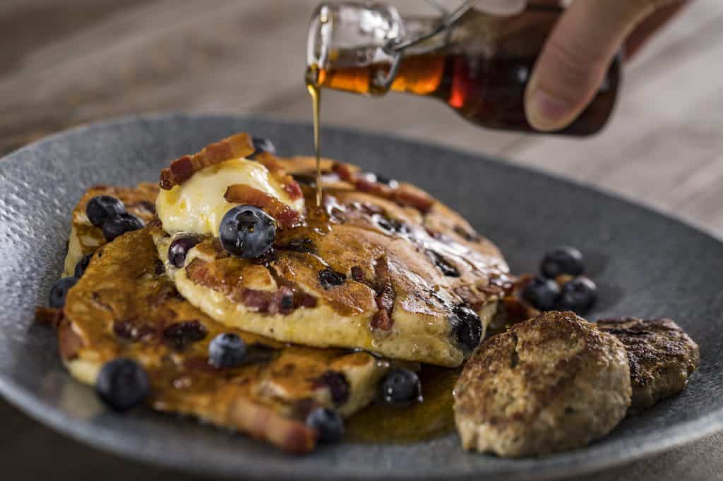 Start your magical morning at Walt Disney World with a plate of fluffy pancakes stacked high, topped with juicy blueberries, crispy bacon strips, and melting butter, all drizzled with golden syrup right before your eyes. This Foodie Guide highlight comes complete with hearty sausage patties on the side—an enchanting breakfast sure to delight theme park fans of all ages.