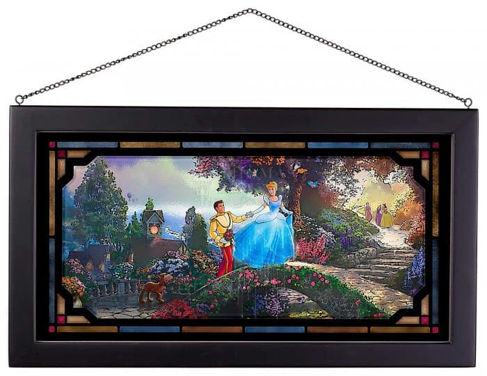 Stained glass artwork featuring a prince and princess from a fairytale kingdom holding hands in an enchanting, colorful garden with a winding path and their playful feline companion nearby, evoking the magical style seen in Disney Parks art displays.
