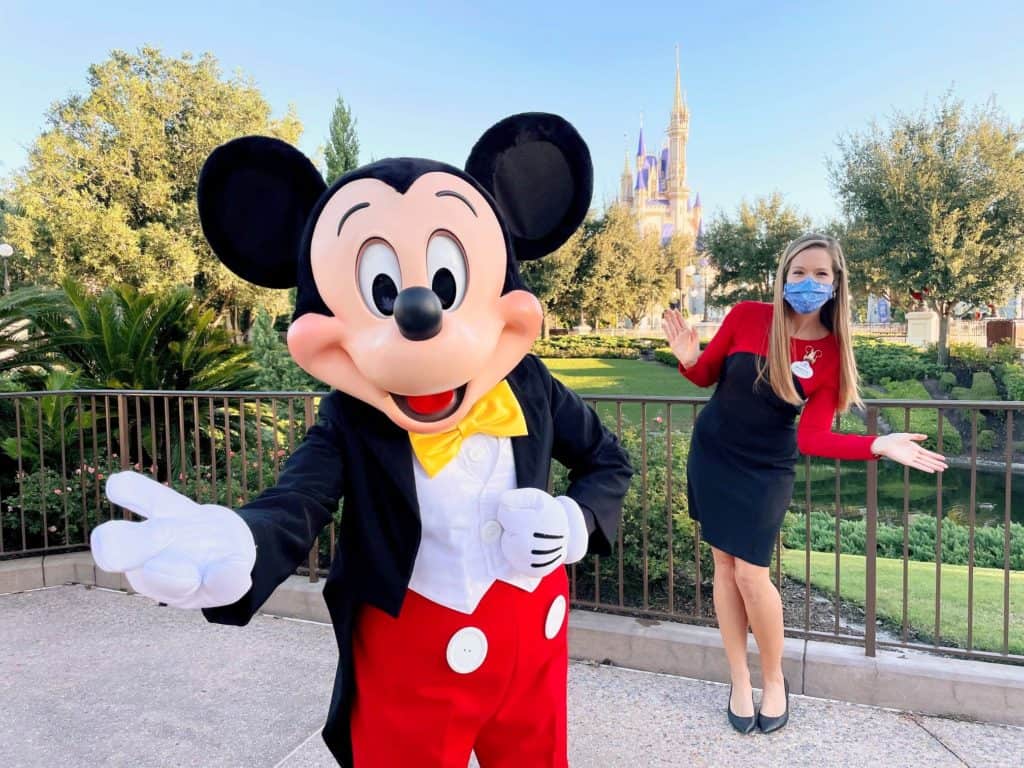 Mickey Mouse greets a guest wearing a face mask in front of the iconic castle at Walt Disney World on a bright, sunny day.