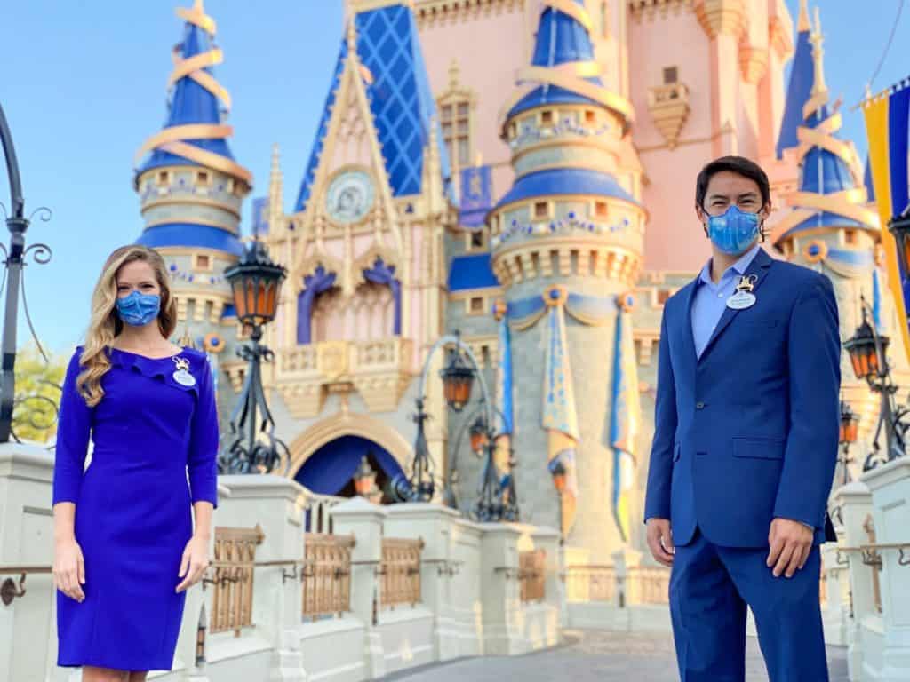 Disney Ambassadors, dressed in signature blue uniforms and masks, pose with bright smiles in front of the iconic Cinderella Castle at Walt Disney World.