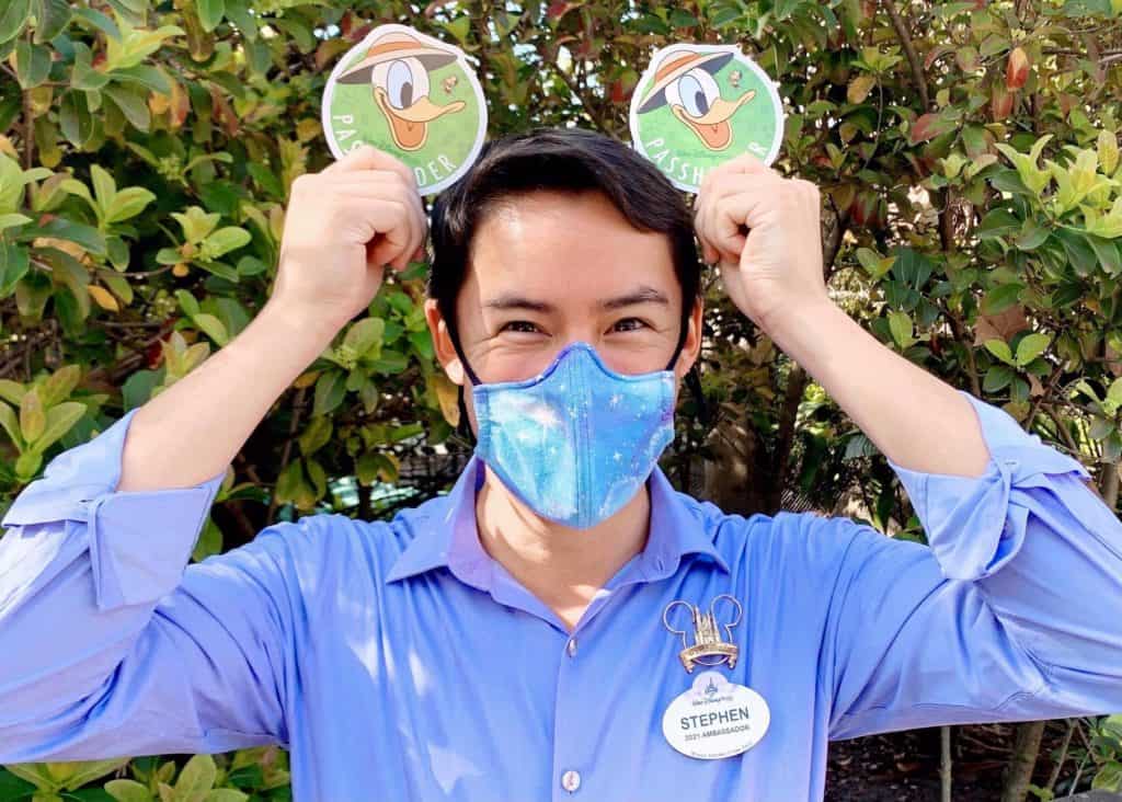 A Disney Ambassador in a blue shirt and mask proudly displays two Donald Duck badges, celebrating their ambassador role amidst lush greenery.