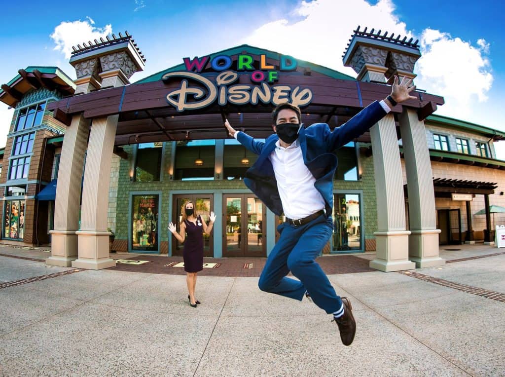 Disney fans, masked up and full of excitement, strike a fun pose outside the World of Disney store—one leaps high with joy, clearly inspired by dreams of joining the legendary Disney Ambassadors who bring Walt Disney World’s magic to life.
