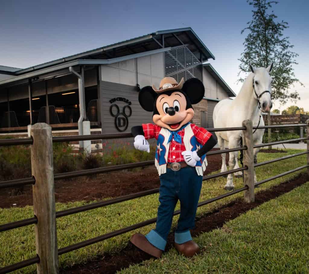Mickey Mouse, dressed as a cowboy, greets guests beside a horse and rustic fence outside the new barn at Disney’s Tri-Circle-D Ranch. A signature Mickey logo is featured on the barn, adding a festive touch for the Mickey anniversary celebration.