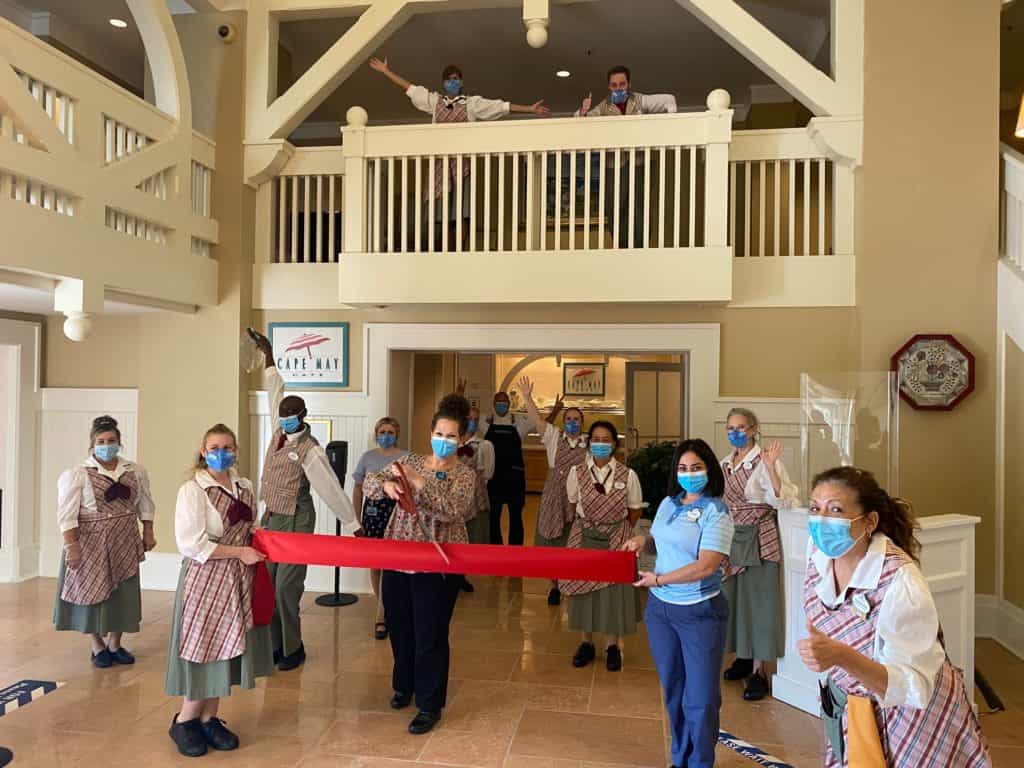 Disney cast members donning face masks proudly hold a red ribbon in the freshly reopened lobby of Cape May Café, marking the celebratory return of this beloved Disney dining spot.