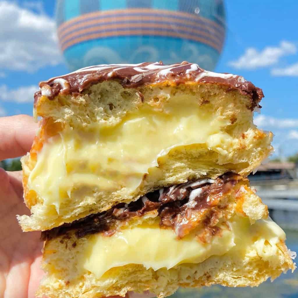 Enjoying a delicious Boston Cream Donut oozing with yellow custard and topped with rich chocolate, perfectly framed by the iconic Aerophile balloon rising over Disney Springs.