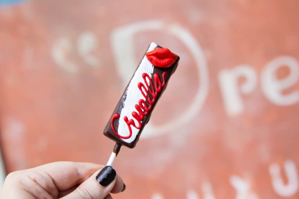 Cruella-inspired chocolate pop with bold red "Cruella" lettering and signature red lips on top, just like the treats found at Disney Springs, held up against a softly blurred background.