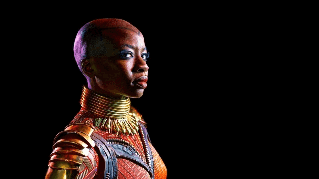 Okoye from Black Panther stands powerfully in signature gold and red armor with a tall neck ring, channeling Danai Gurira’s fierce energy against a dramatic black backdrop, ready to greet guests at the Wakanda attraction.