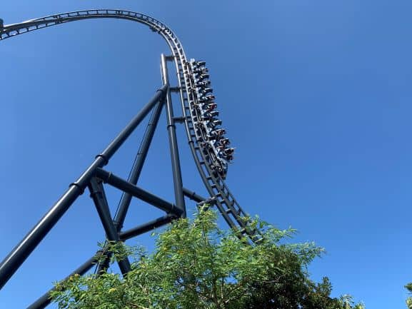 Jurassic World VelociCoaster plunges down its signature steep drop at Universal Orlando Resort, set against a brilliant blue sky with lush trees surrounding the track.
