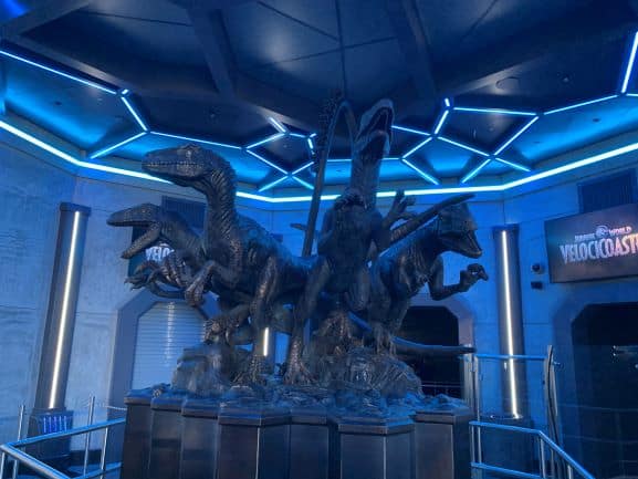 The iconic metallic raptor statue, joined by four dynamic dinosaur figures, stands beneath blue futuristic lights inside Universal Orlando Resort’s VelociCoaster queue, immersing fans in the ride’s intense adventure atmosphere.