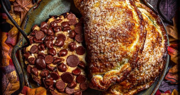 A classic Gideon's Bakehouse chocolate chip cookie and two sugar-dusted Hand Pies are artfully arranged on a rustic plate, surrounded by vibrant autumn leaves, perfectly capturing the warm, festive spirit of Disney Springs.