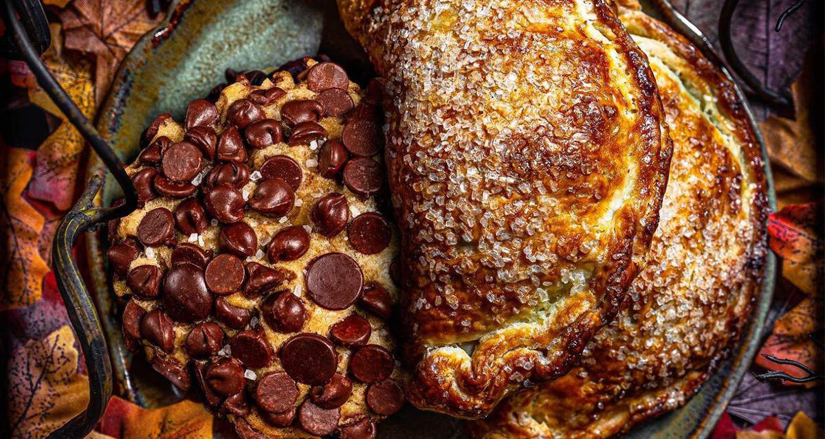 A classic Gideon's Bakehouse chocolate chip cookie and two sugar-dusted Hand Pies are artfully arranged on a rustic plate, surrounded by vibrant autumn leaves, perfectly capturing the warm, festive spirit of Disney Springs.