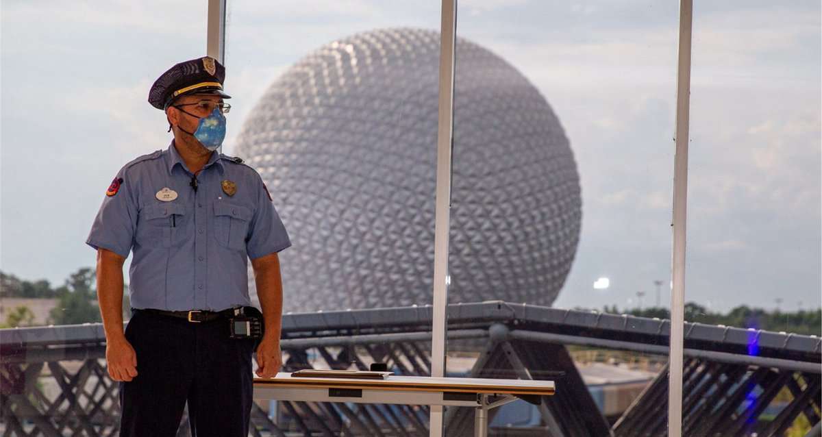 A dedicated Disney Cast Member in official uniform stands inside a park building, with the iconic Spaceship Earth sphere at EPCOT shining through the window behind him—a true everyday hero celebrated with the Disney Heroes Award for saving a child.