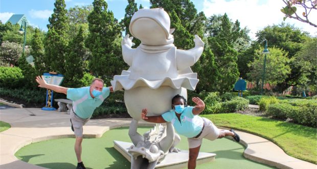 Guests in face masks strike playful poses next to a quirky statue at Fantasia Gardens Miniature Golf, surrounded by vibrant greenery—celebrating 25 years of Disney Cast Members creating magical moments.