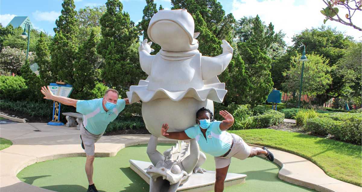 Guests in face masks strike playful poses next to a quirky statue at Fantasia Gardens Miniature Golf, surrounded by vibrant greenery—celebrating 25 years of Disney Cast Members creating magical moments.