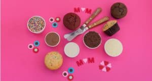 Cupcakes topped with colorful sprinkles and surrounded by baking tools rest on a pink background, featuring "MOM" letter decorations and hearts—capturing the playful, celebratory spirit of Disney Springs and inviting fans to create their own magical Mother’s Day cupcake experience at home.