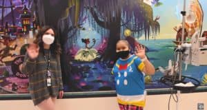 Two team members in masks wave beside medical equipment, set against a vibrant Disney mural inspired by legendary Imagineering, bringing signature Disney magic to brighten the Children’s Hospital environment.