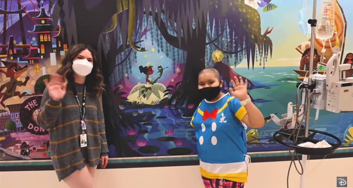 Two team members in masks wave beside medical equipment, set against a vibrant Disney mural inspired by legendary Imagineering, bringing signature Disney magic to brighten the Children’s Hospital environment.