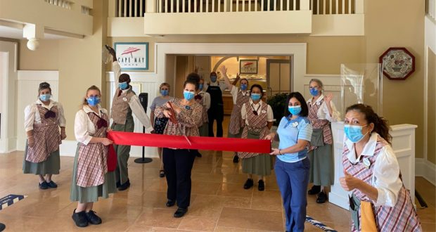 Cast members and staff wearing masks kick off the reopening celebration at Disney’s Cape May Café, holding a red ribbon inside the cheerful, beach-themed restaurant.