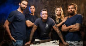 The adventurous Shipwreck Hunters Australia team gathers around a table strewn with maps and tools, confidently preparing for their next thrilling quest in a dim, industrial-themed setting—capturing the excitement and spirit of discovery showcased on Disney+.