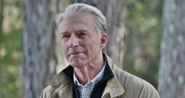Old Man Steve from Avengers: Endgame poses outdoors in front of lush trees, dressed in his iconic beige jacket and checked shirt, just like when he appeared at the end of the film.