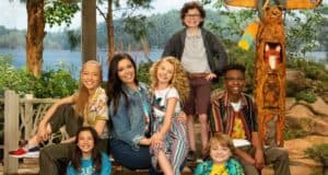 Six smiling kids gather for a photo beside a sparkling lake at an adventure-themed camp, surrounded by lush trees and a carved wooden totem pole, evoking the playful atmosphere of Disney’s Bunk’D Summer Camp as seen in Season 4.