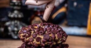 A wooden puppet hand, like something out of a Disney attraction, presents a tempting chocolate chip cookie above a stack of more oversized cookies on a table—evoking the signature treats fans line up for at Gideon's Bakehouse in Disney Springs.